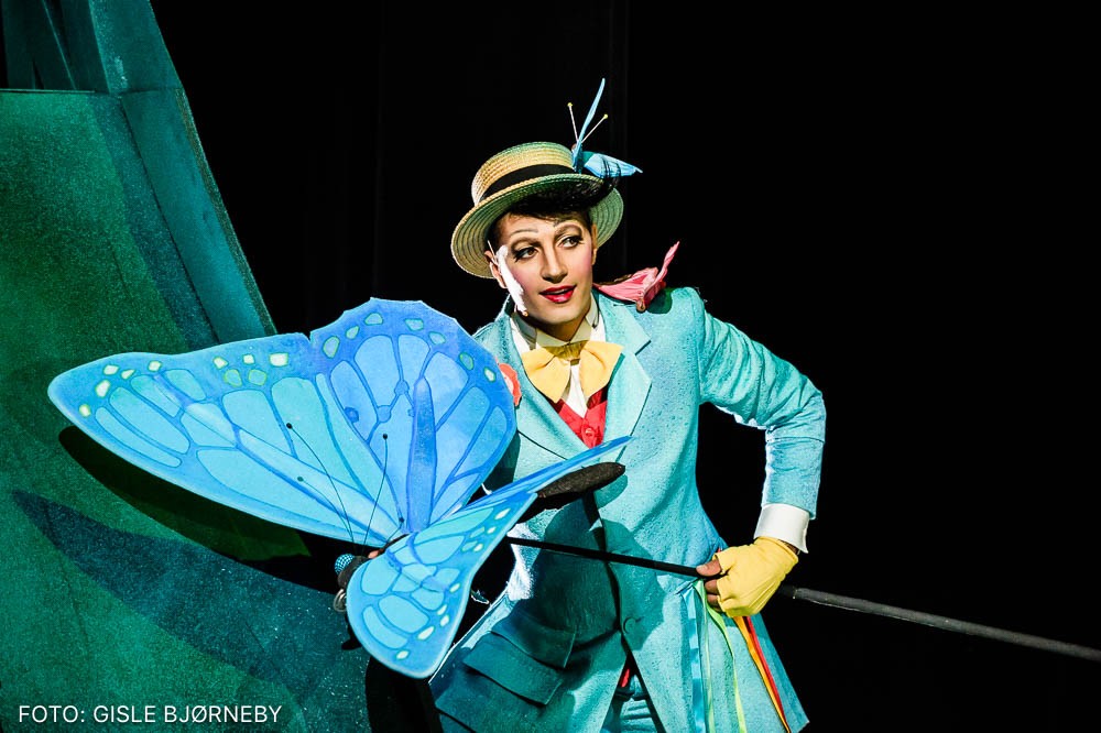 En skuespiller i et blått kostyme og gul hatt som holder en stor sommerfugl festet på en stang