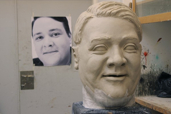 A big head is being modelled and casted. There is a enlarged photograph of the actor in the background